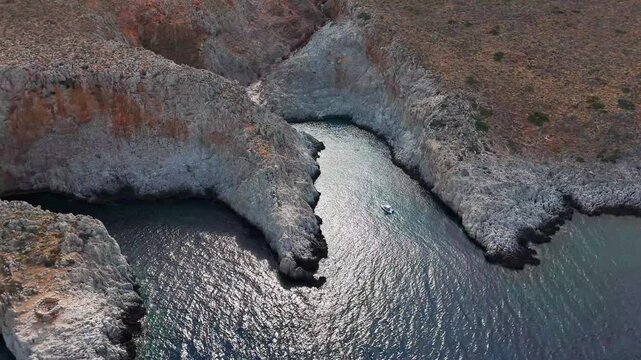 Rugged cliffs divide two narrow coves at Seitan Limania, Crete, Greece, while sunlight reflects across rippled water and the camera passes high above the rocky coast.