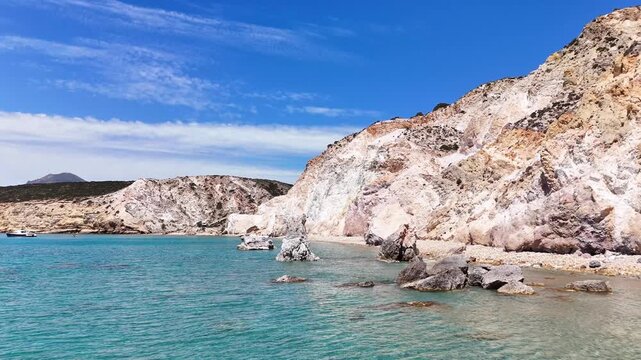 Turquoise water laps beneath pale multicolored cliffs at Paralia Firiplaka on Milos, Greece, with a rocky shoreline, distant boats and thin clouds under bright sun.
