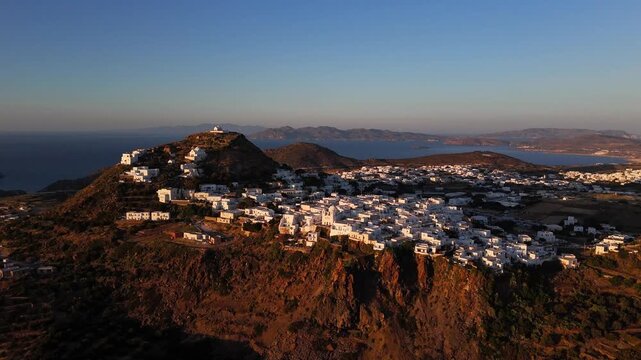 White buildings across Plaka on Milos, Greece, catch the last warm light as the camera looks over the hilltop village, dark rocky slopes and distant Aegean islands.