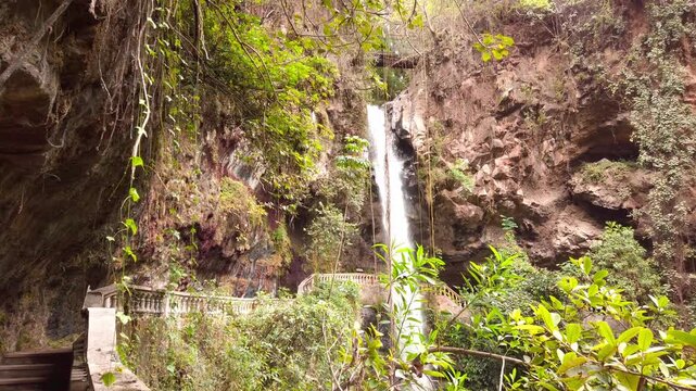 Waterfall cascading through narrow rocky canyon, surrounded by trees, vines, green plants and weathered rock walls with a stone railing near the base in Mexico