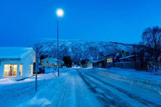 View of a snow-covered street at blue hour with a lit wooden house and a mountain in the background in Norway.