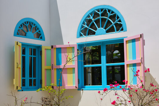 Colorful seaside house facade with turquoise windows, arched glass fanlights, open pastel shutters, and flowering bougainvillea creating cheerful exterior