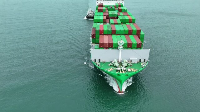 green container ship fully loaded with colorful cargo escorted by two tugboats in the sea. Global maritime logistics, international shipping trade, and freight transport.