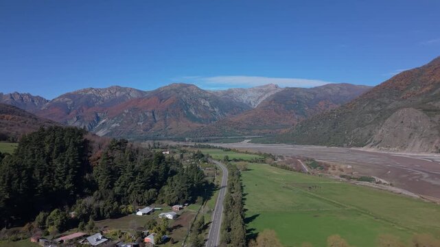 Orbiting pass circling a small rural settlement beside green farmland and a wide braided riverbed framed by jagged snow dusted autumn colored mountains