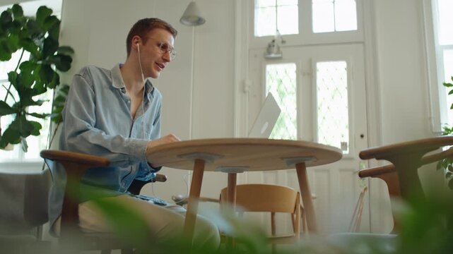 Man joins a laptop video call at a modern workspace table, using an earbud for remote work, online communication, freelance tasks, and a flexible meeting setup.