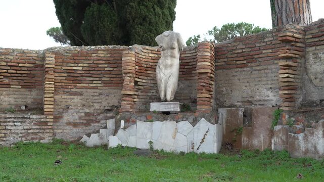 Weathered headless Roman marble torso stands against an old brick wall in Ostia Antica, Italy.