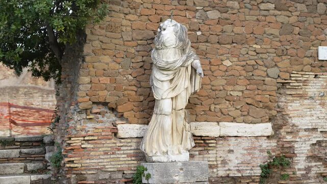 Weathered headless Roman marble statue stands against a rough brick wall, framed by stone steps and a leafy tree in Ostia Antica.