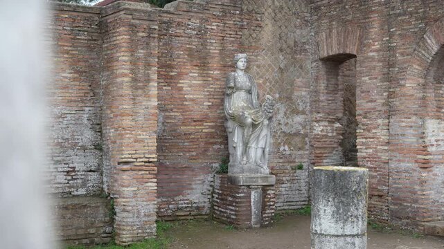 Female figure holding a cornucopia stands in a recessed brick niche with a worn column fragment in the foreground at Ostia Antica.