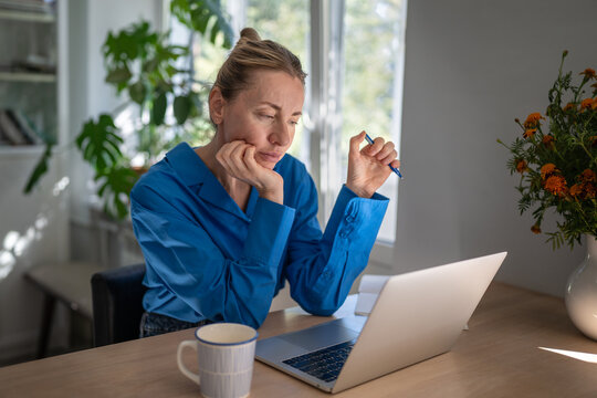 Sleepy pensive businesswoman working on laptop from home sitting at table staring at screen, thoughtful female resting chin on hand at desk in kitchen while looking at computer, tiredness and boredom.