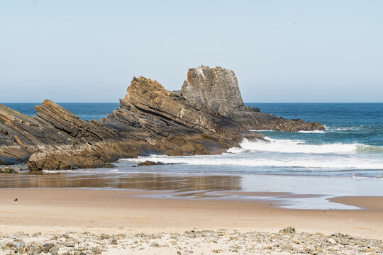 View of dramatic layered rock formations on a sandy beach along the Atlantic coastline under a clear blue sky in Aljezur, Faro District, Portugal.