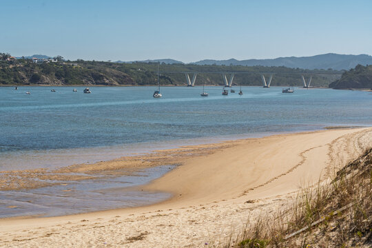 View of the Mira River estuary with sailboats anchored in calm water, a modern bridge in the background, and sandy banks, Vila Nova de Milfontes, Beja District, Portugal.