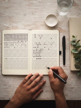 Close up top view of hands writing in a habit tracker notebook filled with dot grid checklists on a wooden table.