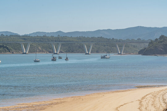 View of the bridge over the Mira River with sailboats anchored in the calm water near the sandy beach in Vila Nova de Milfontes, Beja District, Portugal.
