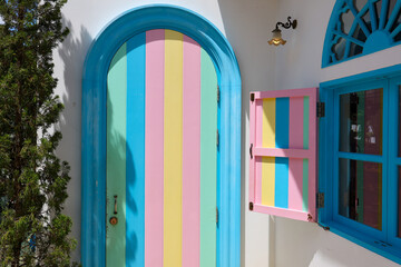 Obraz premium Turquoise arched door and colorful shutter on white Mediterranean house exterior create cheerful geometric facade with vivid pastel stripes and strong sunlight