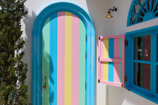 Turquoise arched door and colorful shutter on white Mediterranean house exterior create cheerful geometric facade with vivid pastel stripes and strong sunlight