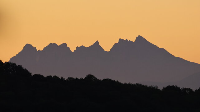 View of the Dents du Midi mountain range silhouetted against a golden sunrise sky above a dark treeline in Val-d'Illiez, Valais, Switzerland.