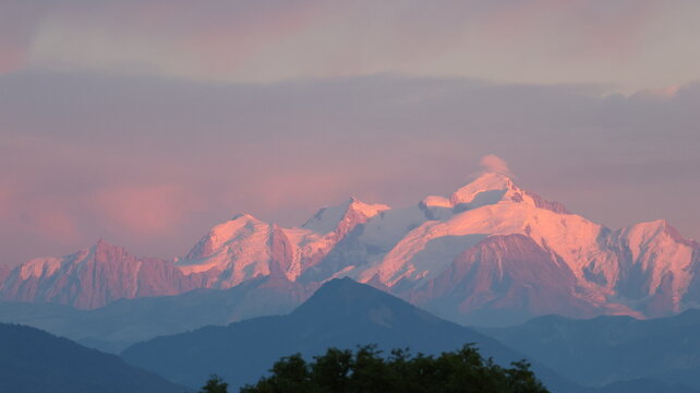 View of the Mont Blanc mountain range illuminated by warm pink sunset light, with snow-capped peaks rising above dark foothills in Chamonix, Auvergne-Rhone-Alpes, France.