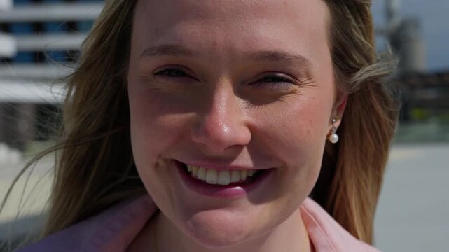 Close portrait of a cheerful young White blonde woman touching her hair, laughing and looking directly at the camera in bright sunlight.