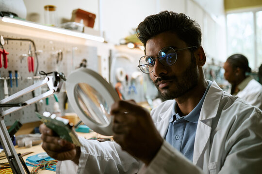Indian young engineer examining circuit board with magnifying glass in laboratory. Image suitable for engineering campaigns, robotics research, and electronic hardware development