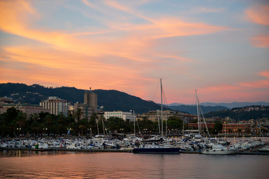 View of the harbor in La Spezia with numerous sailboats and yachts moored at the docks against a vibrant sunset sky over the coastal hills of La Spezia, Liguria, Italy.