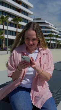 Vertical video of a smiling young blonde woman sitting outdoors and reading content on her smartphone in a sunny modern urban setting.