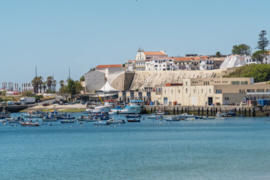 View of the harbor in Sines with fishing boats moored in calm blue water, set against historic white buildings on a cliff under a clear sky in Sines, Setubal, Portugal.