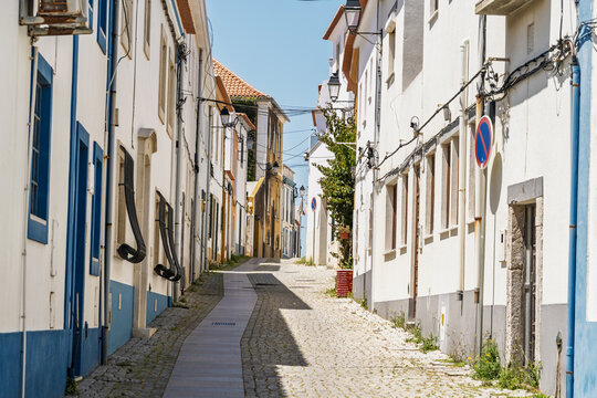 View of a narrow cobblestone street in the historic old town of Sines, lined with traditional white buildings featuring blue trim, under a clear blue sky in Sines, Setubal, Portugal.