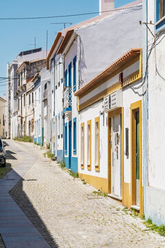 View of a narrow cobblestone street lined with traditional white-washed buildings featuring blue and yellow trim in the historic town of Sines, Setubal, Portugal.