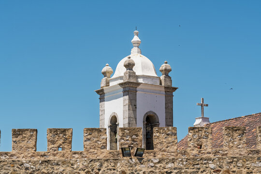 View of the historic church bell tower rising behind a stone crenellated wall under a clear blue sky in Sines, Setubal, Portugal.