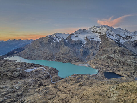 View of Engadin valley featuring the turquoise waters of Lago Bianco surrounded by snow-capped mountain peaks under a soft sunset sky in Grisons, Switzerland.