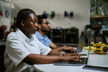 Fototapeta premium African American young woman typing on laptop in engineering workshop alongside colleague. Useful for IT software development branding and STEM education