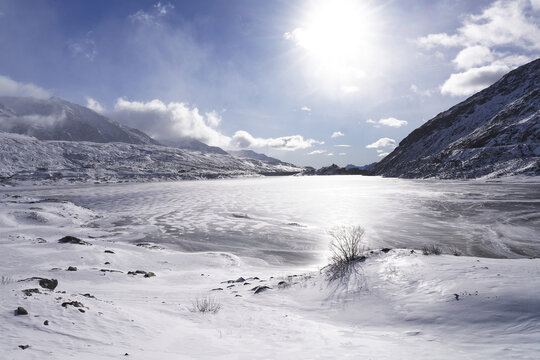 View of the frozen Lake Montespluga surrounded by snow-covered mountains under a bright winter sun in Montespluga, Lombardy, Italy.