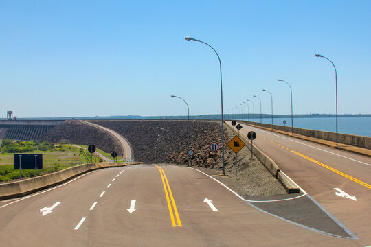 View of Itaipu Dam with a paved road leading across the massive stone embankment under a clear blue sky in Itaipulandia, Parana, Brazil.