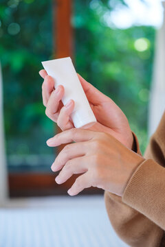 Vertical closeup image of a woman holding a white tube and applying cream on hand