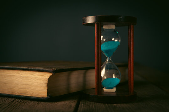 Hourglass and old book on wooden table, vintage color tone.