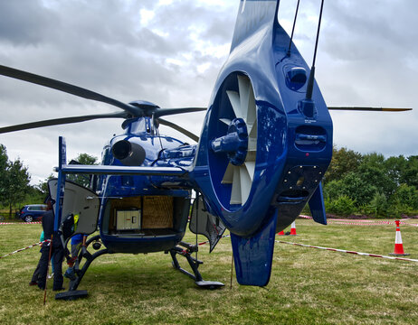 Blue helicopter standing on grass field, preparing for flight under cloudy sky, rear view of tail rotor