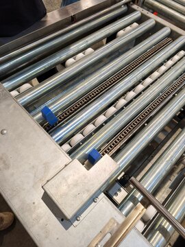A high-angle shot of an industrial roller conveyor system, featuring stainless steel rollers, drive chains, guidance wheels, and metallic frames used for material handling and automated production lin