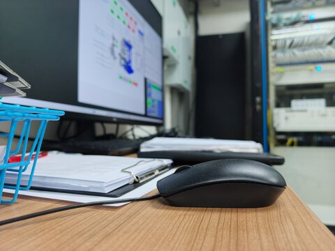A low-angle desktop view in an engineering or server control room, showing a black computer mouse on a wooden desk, clipboard with documents, and a blurred computer monitor displaying monitoring softw