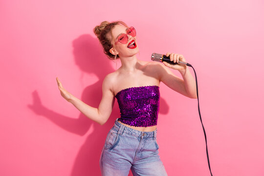 Young woman singing with a microphone, wearing glamorous sequined outfit against pink background, expressing joy and energy