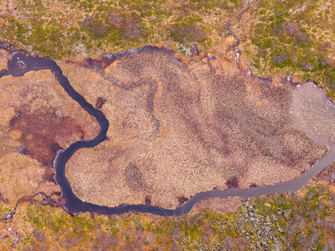 Aerial view of a winding river flowing through a marshy tundra landscape with mossy ground, scattered rocks, and autumn vegetation in Fjord, More og Romsdal, Norway.
