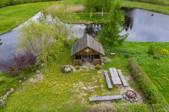 Aerial view of a rustic wooden gazebo and picnic area with a fire pit situated on a lush green lawn filled with yellow dandelions beside a tranquil pond in Latvija.