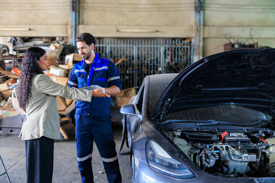 Male auto mechanic in a blue uniform holding a clipboard and explaining the car repair estimate to a female customer next to a car with an open hood in an auto repair shop.