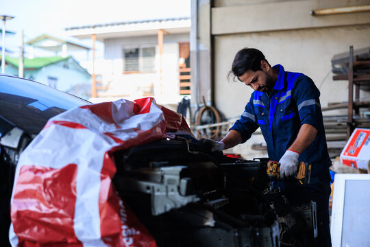 Male auto mechanic in a blue uniform repairing a car engine with a cordless power tool at an outdoor auto repair shop.