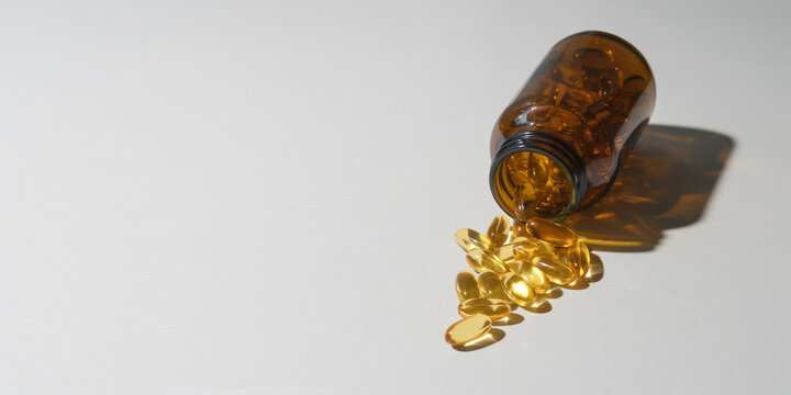 Amber supplement bottle spills golden softgel capsules across white background under hard light with wide copy space for healthcare.