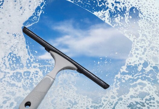 A squeegee cleaning a soapy glass window with a clear blue sky and clouds in the background