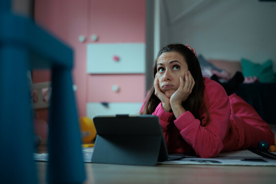 Bored Young Female Resting Chin In Hands While Working On Tablet. Exhausted lady wearing fuchsia lounge wear looks up tiredly near her portable screen