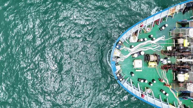 multiple ship crew members in bright safety orange working with massive ropes and heavy machinery on the vibrant green deck of a large vessel's bow, with blue water surrounding
