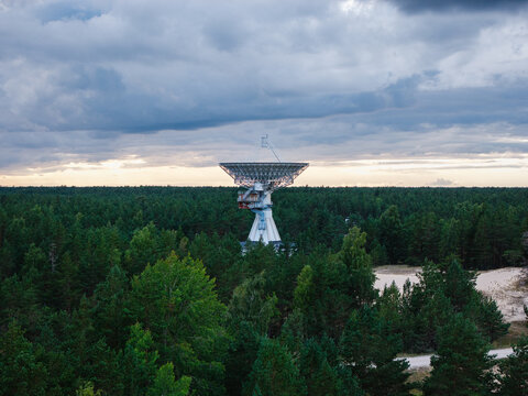 Aerial view of the Irbene radio telescope with its massive parabolic dish rising above the surrounding dense forest under a cloudy twilight sky in Irbene, Ventspils novads, Latvia.