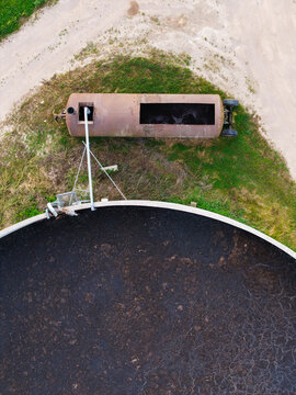 Aerial view of a large circular storage tank filled with dark material next to a rusty cylindrical tank on a grassy field in Virpe, Ventspils novads, Latvia.