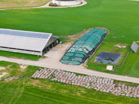 Aerial view of a farm in Virpe featuring a barn with solar panels, a covered silage pit, a small pond, and rows of hay bales in the green fields in Virpe, Ventspils novads, Latvia.